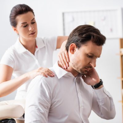 Female massage therapist massaging hurting neck of businessman sitting on massage chair on blurred